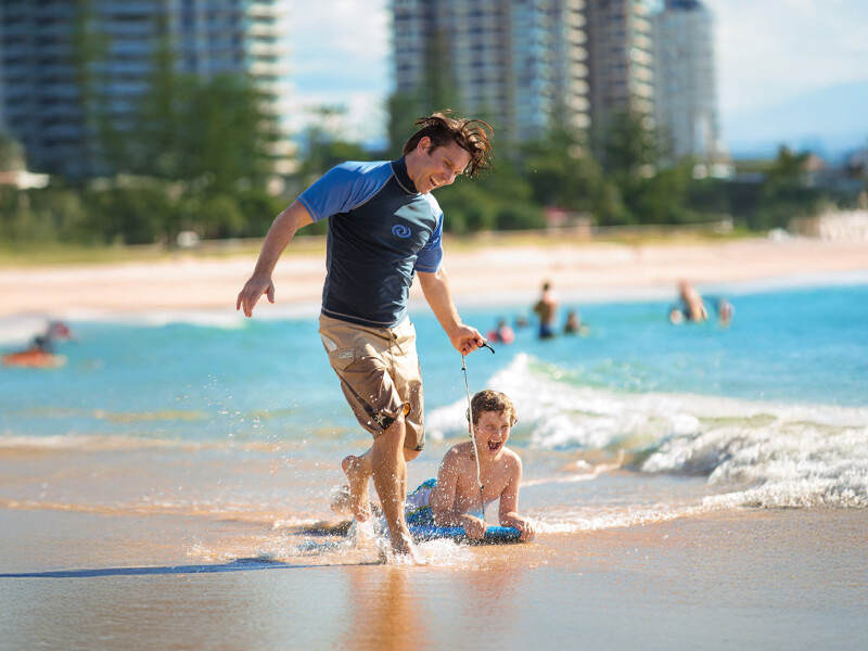 Father pulling his son on a boogie board through shallow waves at Coolangatta Beach, with beachfront buildings in the background.