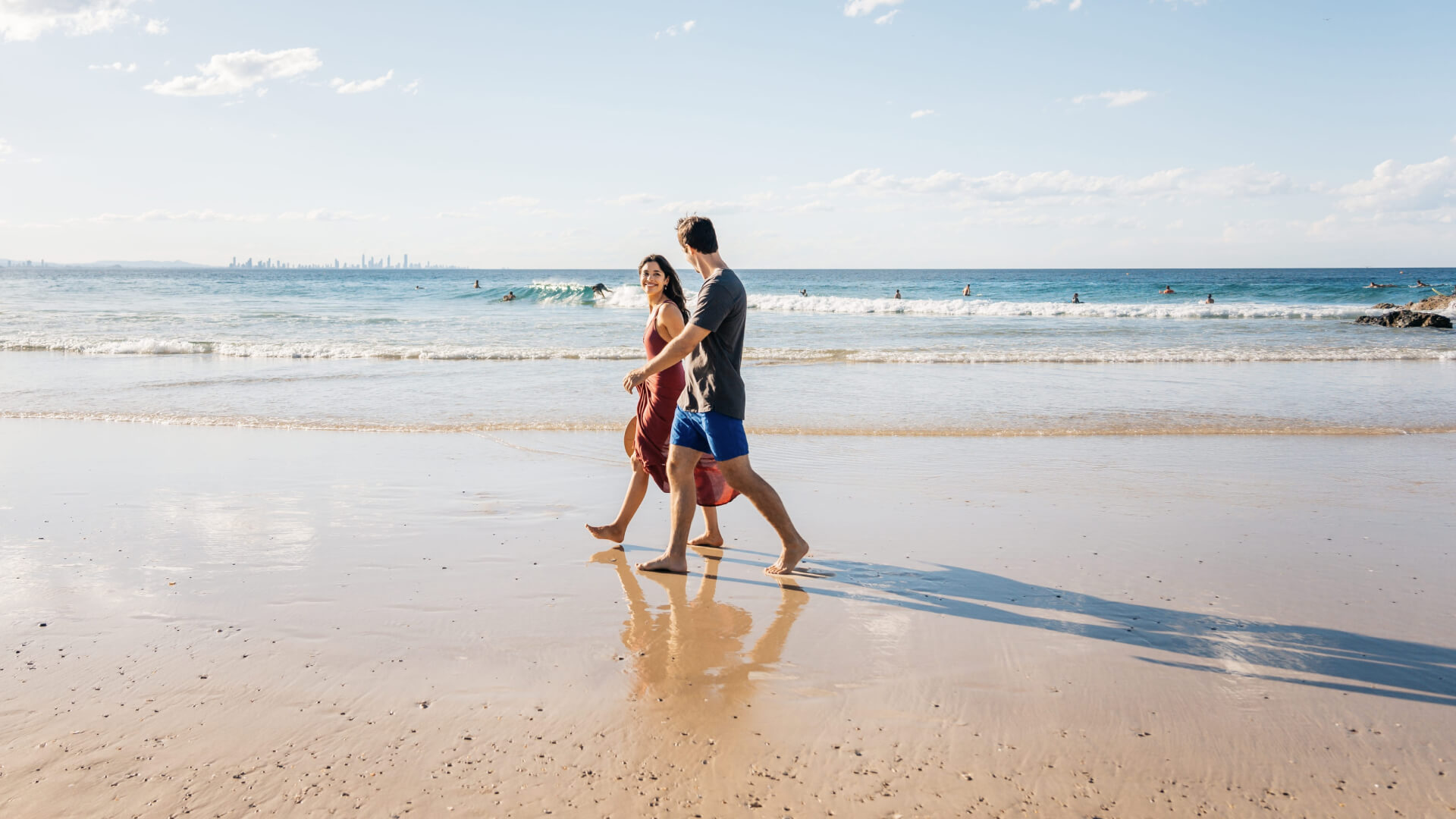 Couple walking barefoot along Coolangatta Beach with gentle waves, surfers in the background, and the Gold Coast skyline visible in the distance under a bright blue sky.