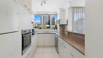 Full kitchen at ULTIQA Beach Haven on Broadbeach with white cabinetry, oven, cooktop, dishwasher, and a window overlooking nearby buildings.