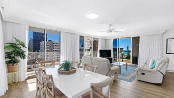 Open-plan dining and living space at ULTIQA Beach Haven on Broadbeach with a white dining table, cream recliners, and large windows with skyline views.