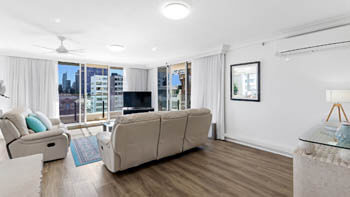 Living area at ULTIQA Beach Haven on Broadbeach with cream recliner lounges, TV, blue rug, and sliding doors leading to the balcony.