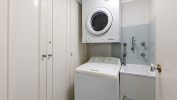 Laundry area with top-loading washing machine, wall-mounted dryer, laundry tub and built-in cupboards at ULTIQA Beach Haven on Broadbeach.