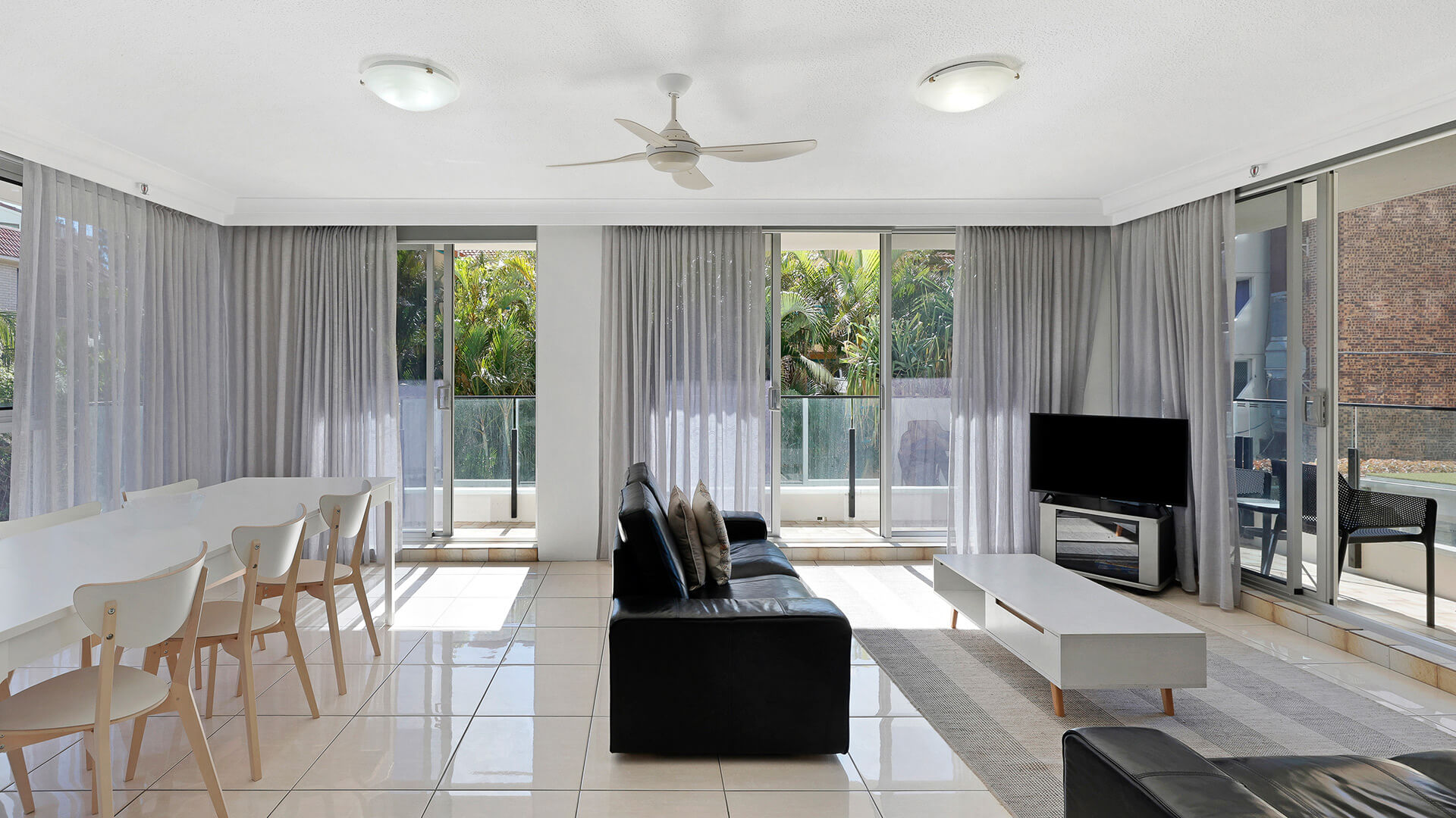 Bright open-plan living & dining area at ULTIQA Beach Haven on Broadbeach, with black leather sofa, dining table and sliding doors to a leafy balcony.
