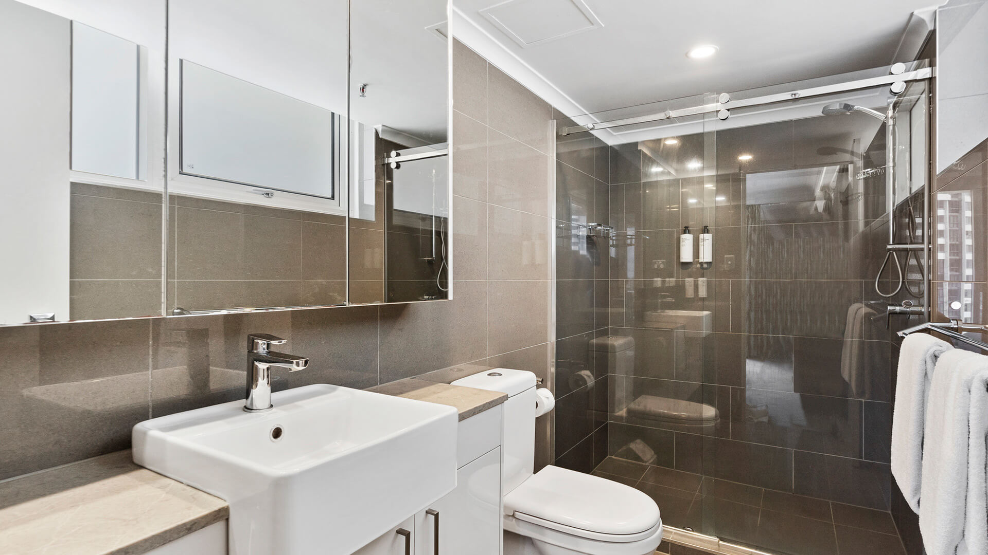 Bathroom with stone tiles, white sink, toilet and a walk-in shower at ULTIQA Beach Haven on Broadbeach.