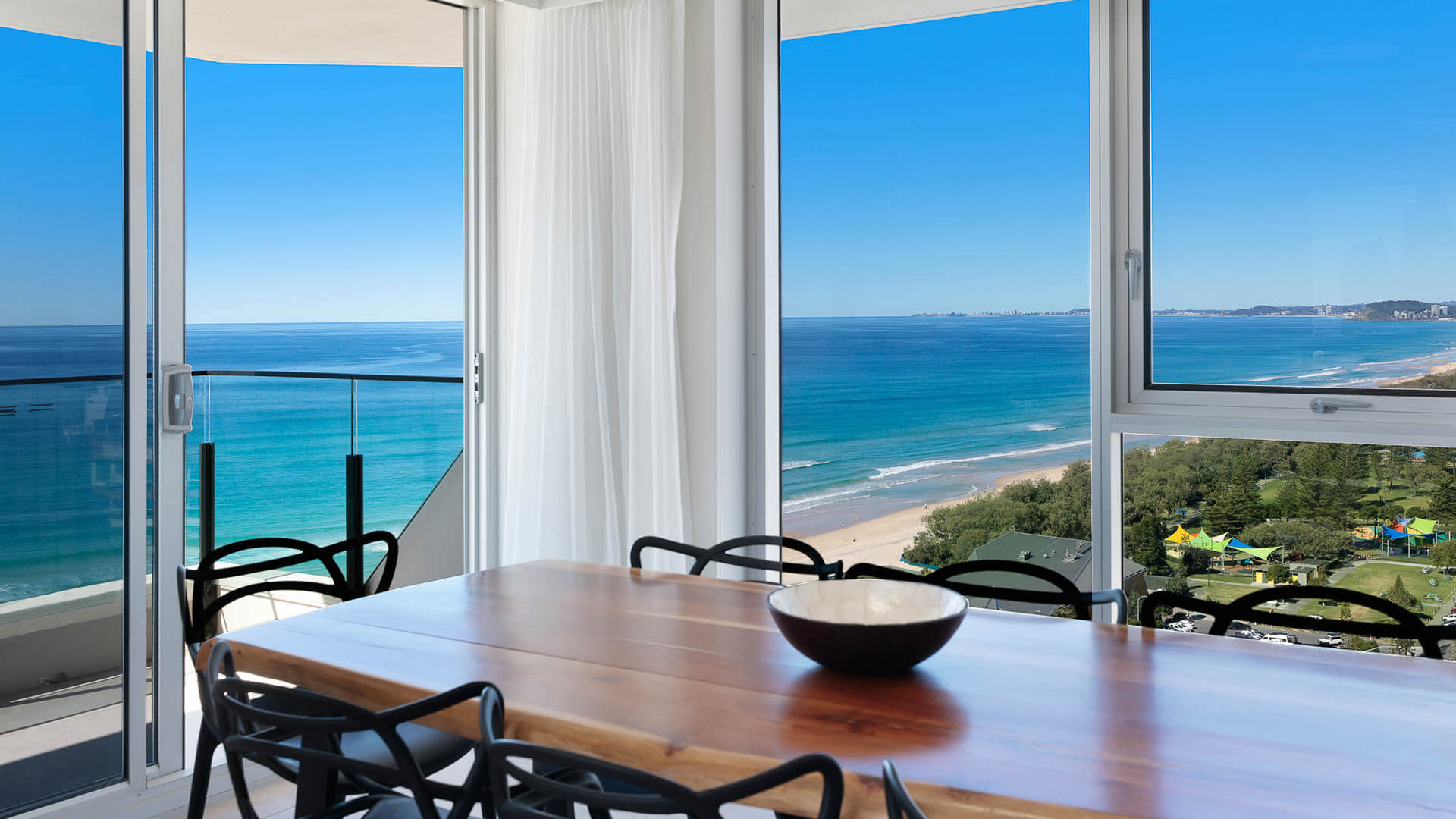 Close-up of the dining area featuring a wooden table, black designer chairs, and sweeping views of the coastline and turquoise ocean. 